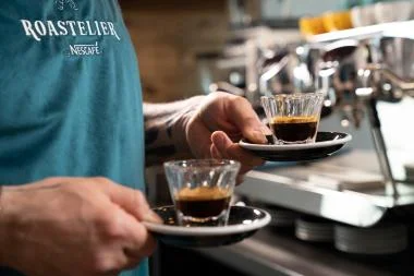 Barista wearing a Nescafé shirt serving two cups of espresso at a coffee machine.