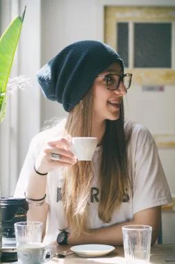A woman wearing a beanie and glasses sits at a table, enjoying a cup of coffee.