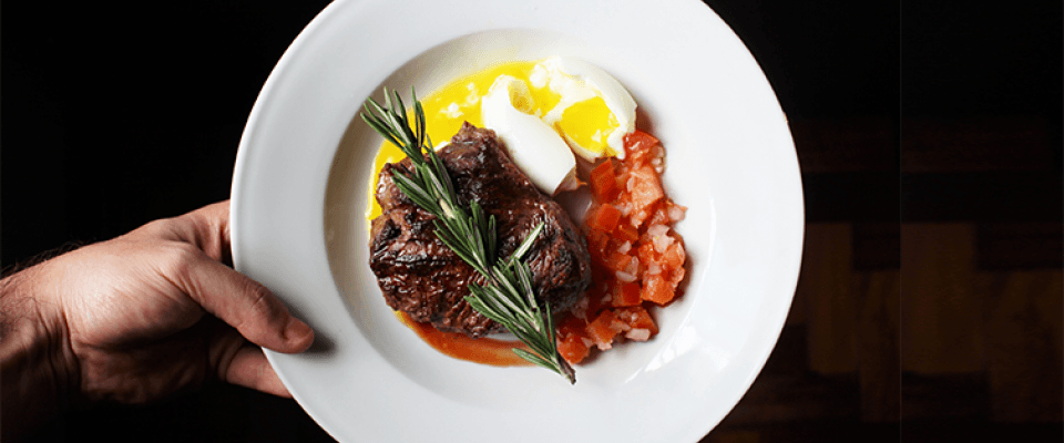 A plate of food featuring cooked meat, fried eggs, and fresh tomatoes arranged neatly.