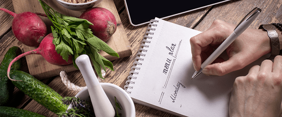 A person writes a menu nutrition plan in a notebook, surrounded by fresh vegetables and herbs.
