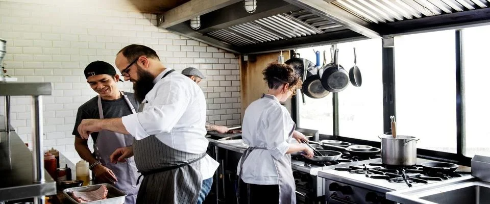 Chefs working together in a busy commercial kitchen, chopping vegetables and cooking on stovetops.