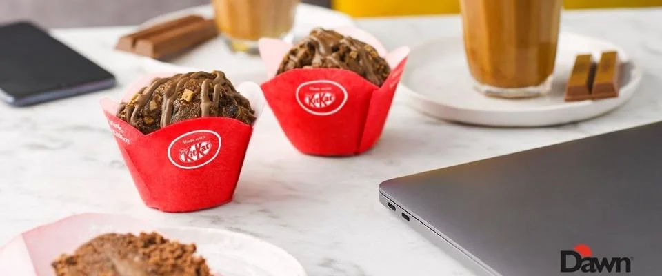 A laptop sits beside a plate of colorful cupcakes and a steaming cup of coffee on a wooden table.