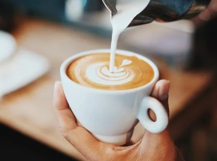 A person pours milk into a steaming cup of coffee, creating a swirl of colors in the drink.