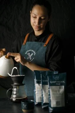 A woman in an apron pours coffee from a carafe into a pot, ready to brew a fresh batch of coffee.