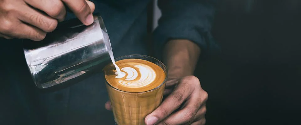 A person pours steaming coffee from a pot into a white cup on a wooden table.