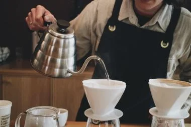A person is pouring steaming coffee from a pot into a white cup on a wooden table.