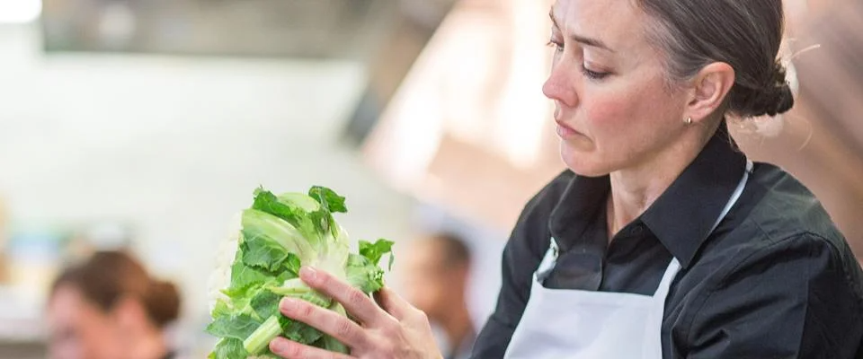 A woman stands in a kitchen, smiling as she holds a fresh salad in her hands.
