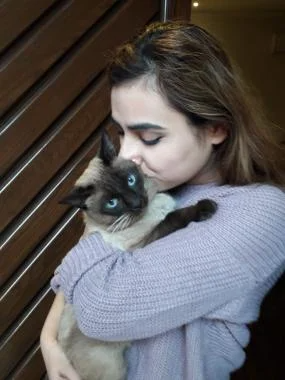 A woman gently holds a Siamese cat in her arms, smiling at the camera.