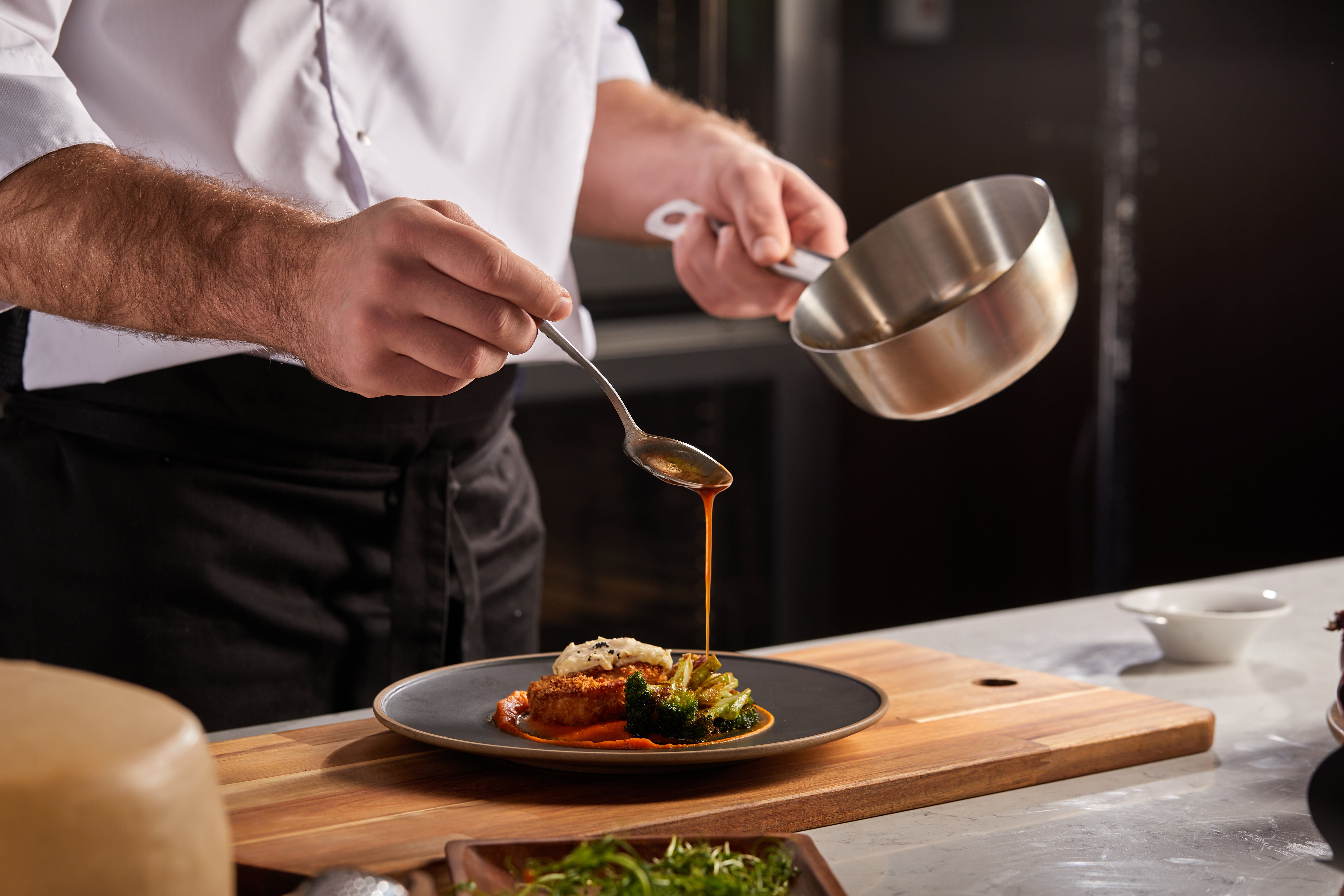 Chef pouring sauce onto plated gourmet dish with vegetables on wooden cutting board.