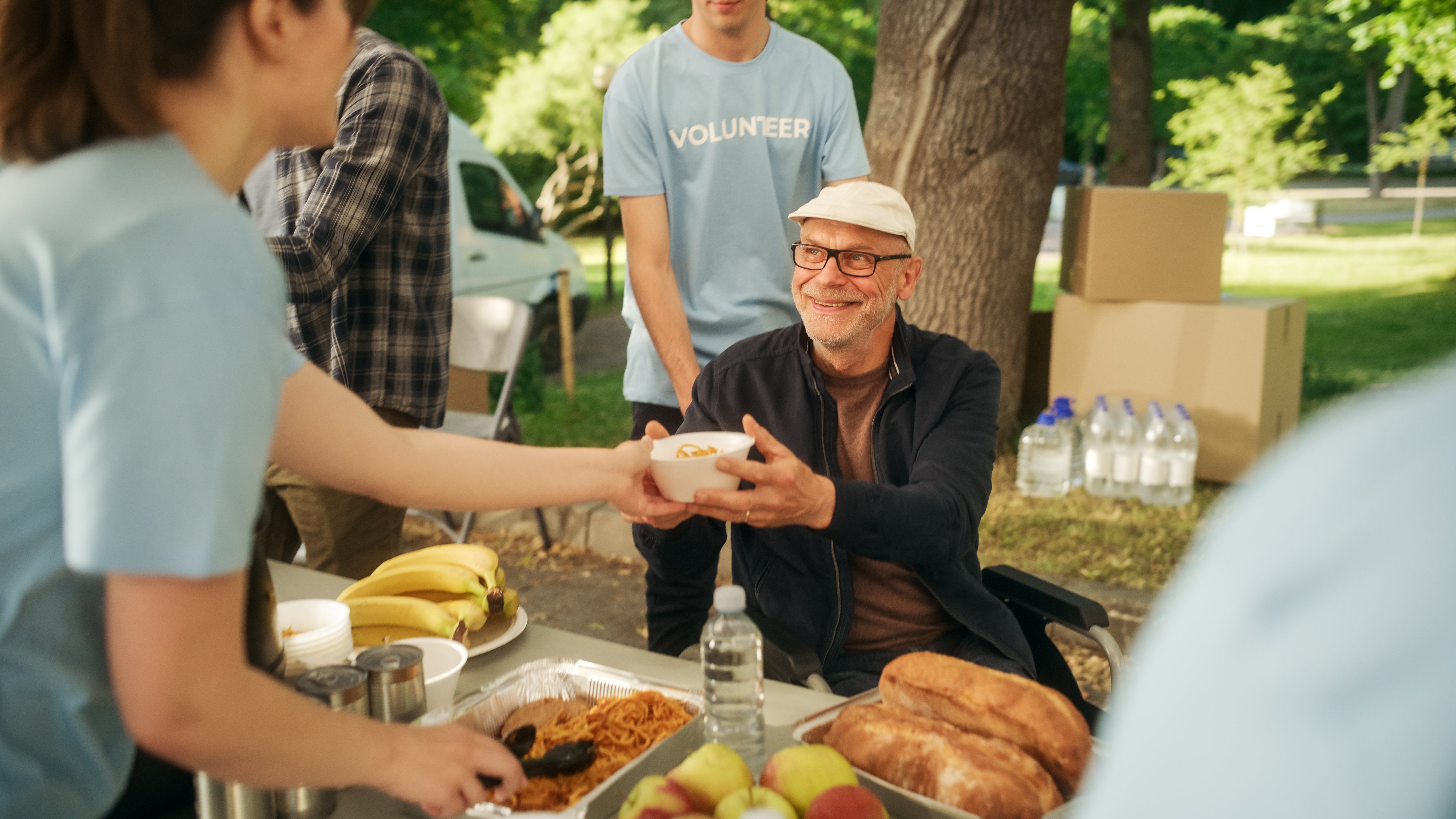 A man hands a plate of food to a woman sitting at a picnic table, surrounded by greenery and outdoor picnic items.