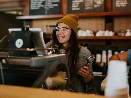 A woman wearing a beanie smiles while serving coffee in a bustling coffee shop.