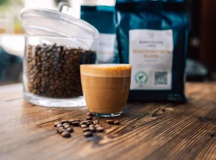 A close-up of coffee beans scattered next to a steaming cup of coffee on a wooden surface.