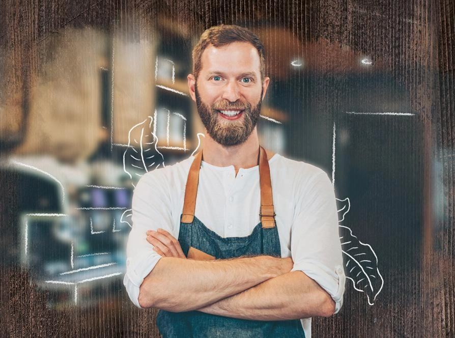 A bearded man in an apron stands in front of a chalkboard, ready to share his knowledge or skills.