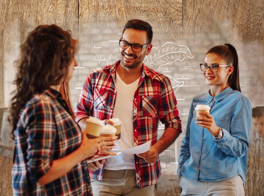 Three people stand in front of a wall, enjoying coffee together and smiling.