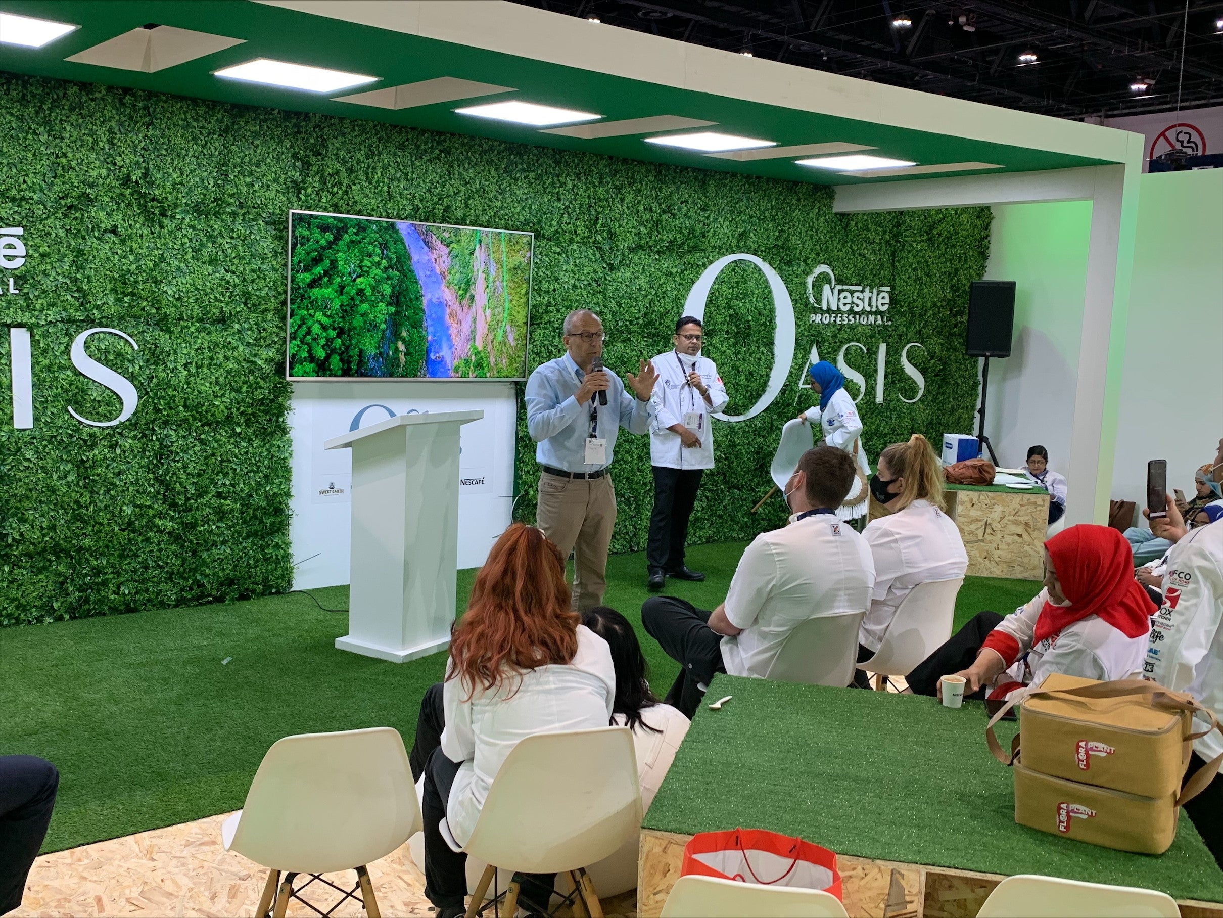 People sitting at tables, chatting and laughing, with a green wall serving as their backdrop.
