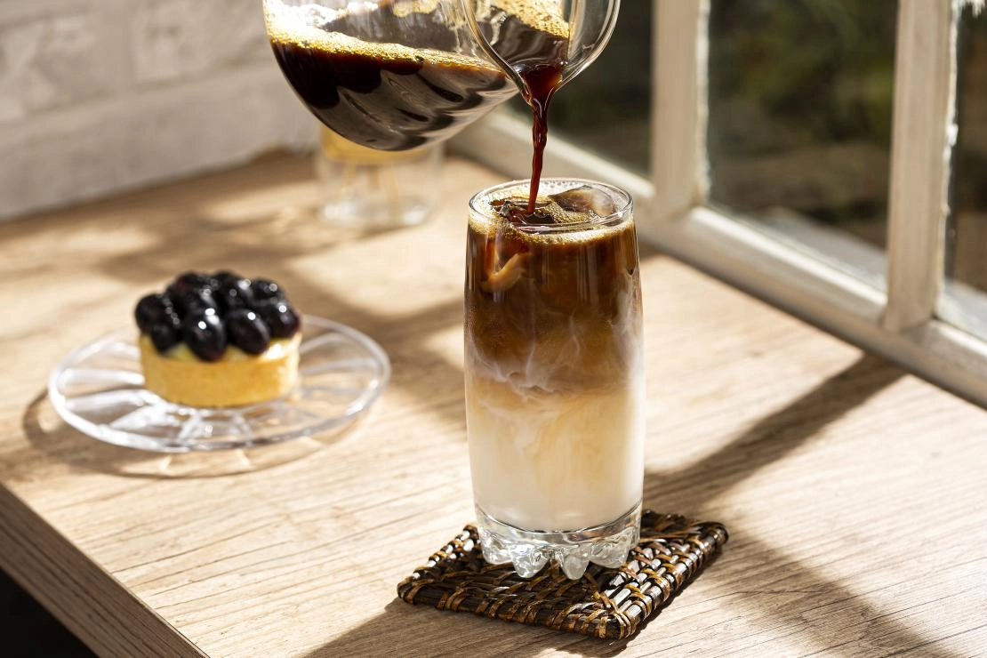 Person pouring hot coffee into a glass filled with ice, creating a refreshing iced coffee beverage.