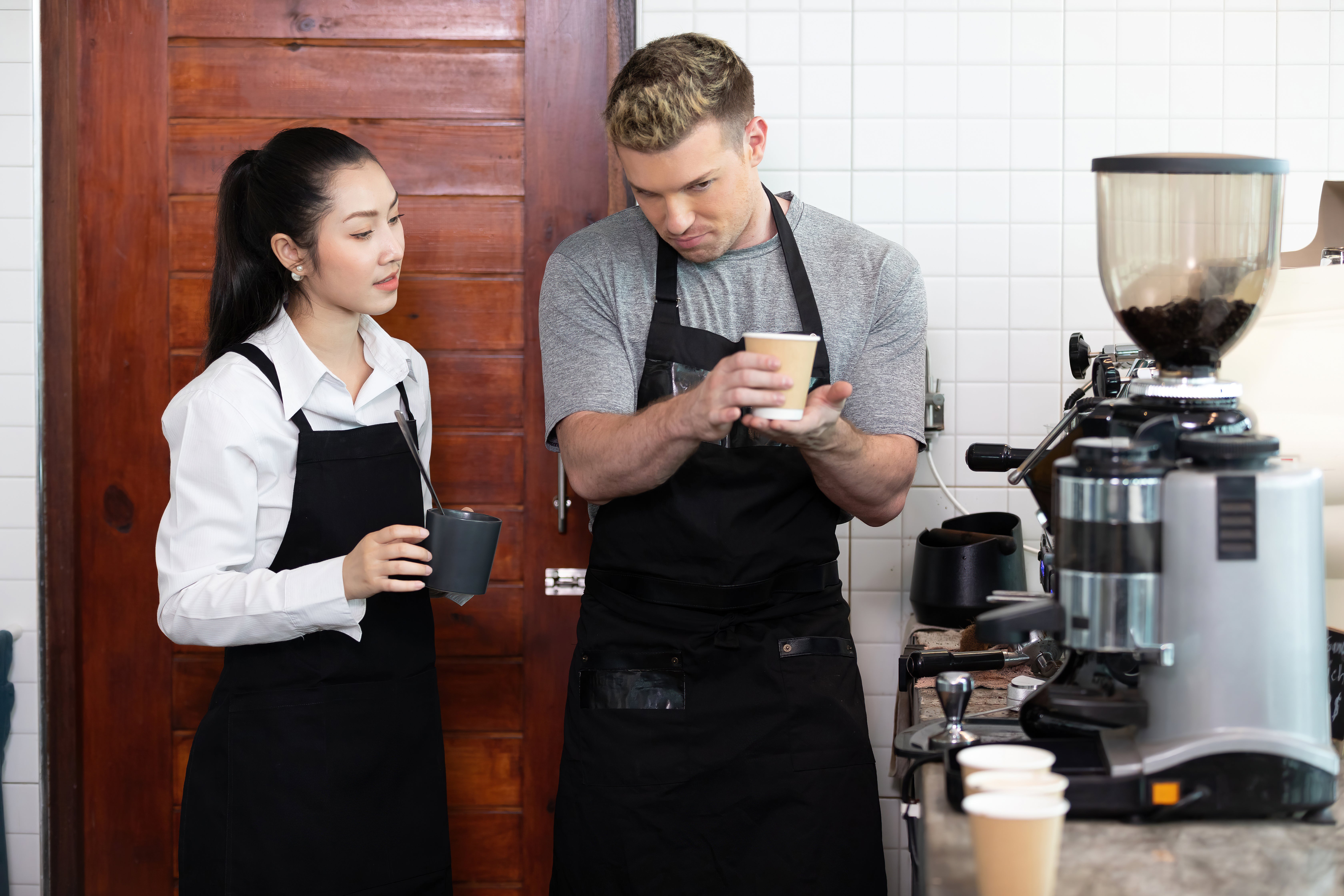 A man and woman stand together, smiling in front of a coffee machine in a cozy kitchen setting.