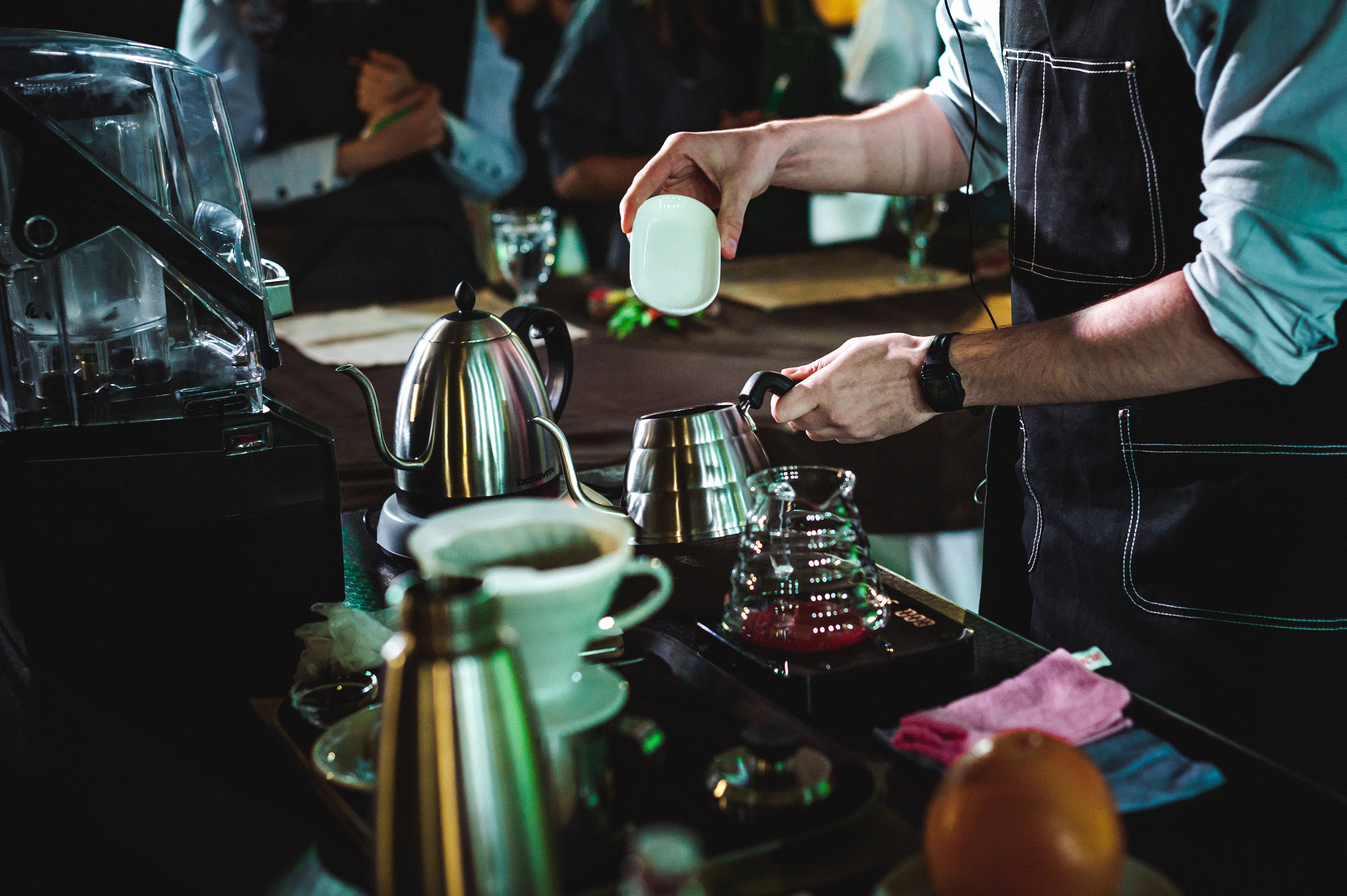 A man in a black apron pours coffee from a pot into a white cup, showcasing a cozy café scene.