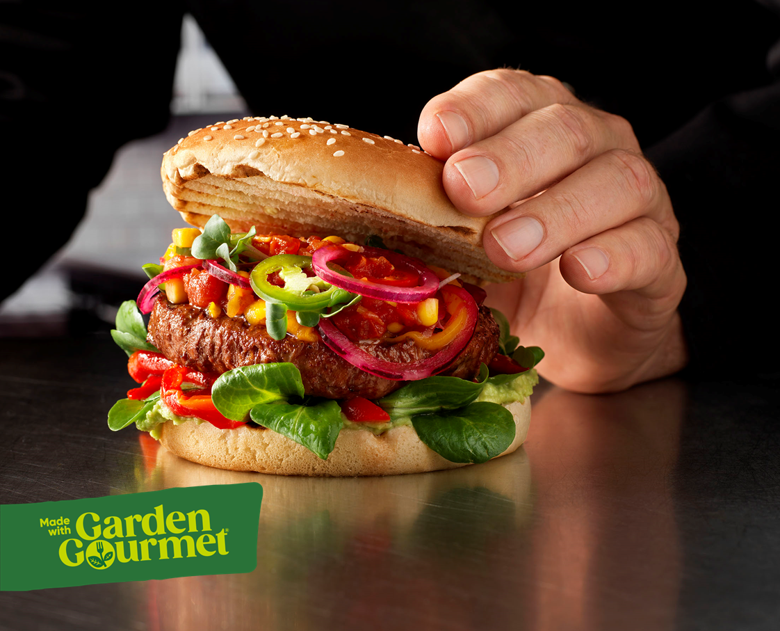 A person holding a burger topped with a fresh green leaf, showcasing a delicious meal option.