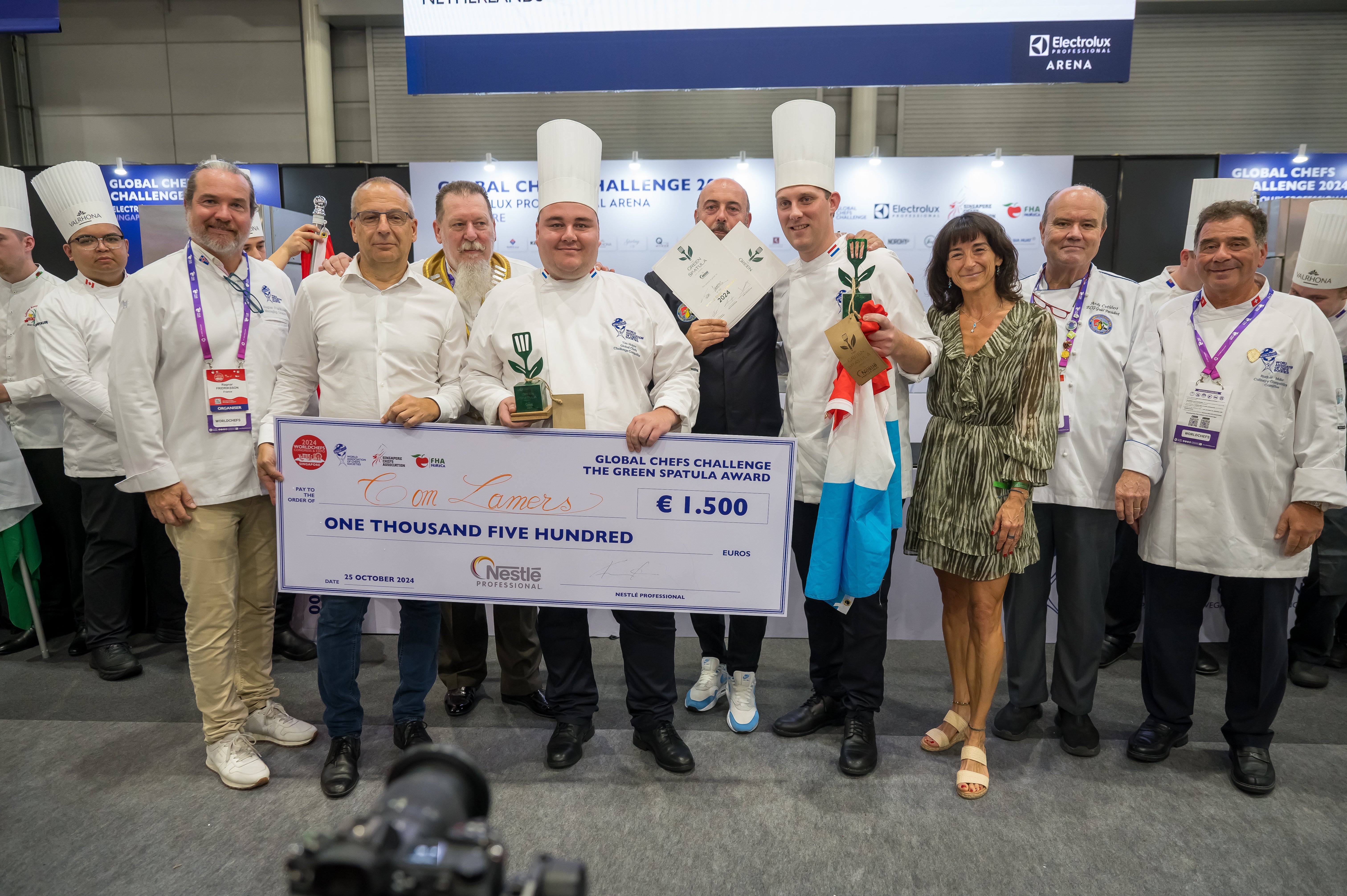 A group of smiling chefs stands together, holding a large check in celebration of their culinary achievement.