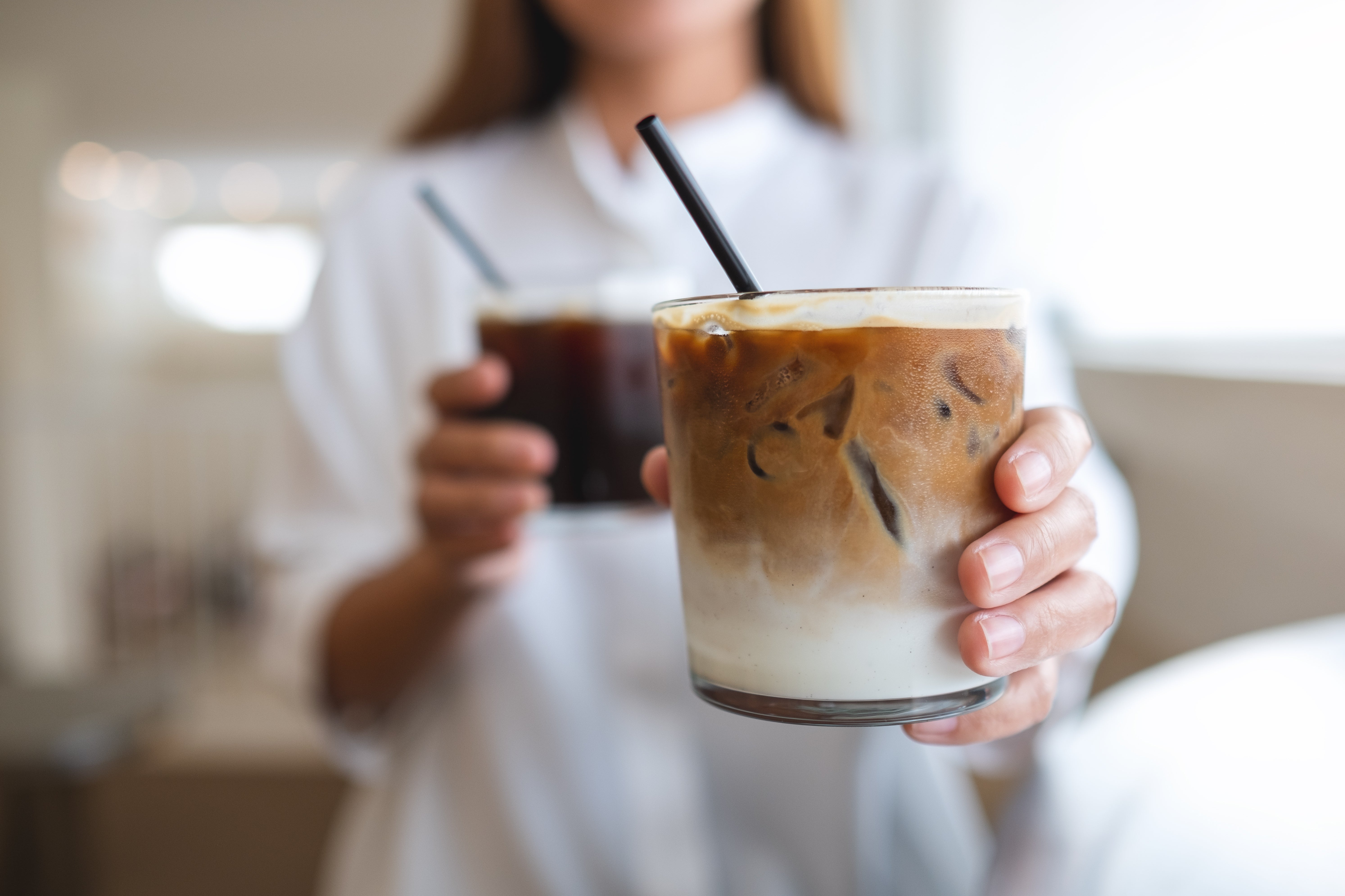 A woman smiling while holding two glasses of iced coffee, showcasing a refreshing beverage choice on a sunny day.