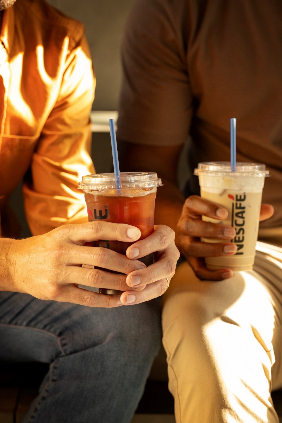 Two people sit on a bench, each holding a Nescafé drink and enjoying a casual conversation.