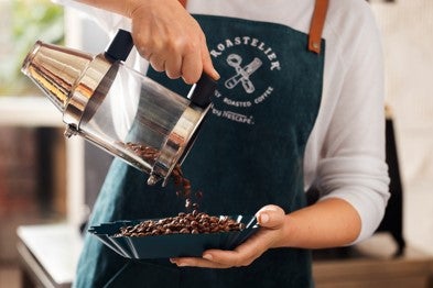 A woman in an apron pours coffee from a pot into a bowl, focusing intently on her task.