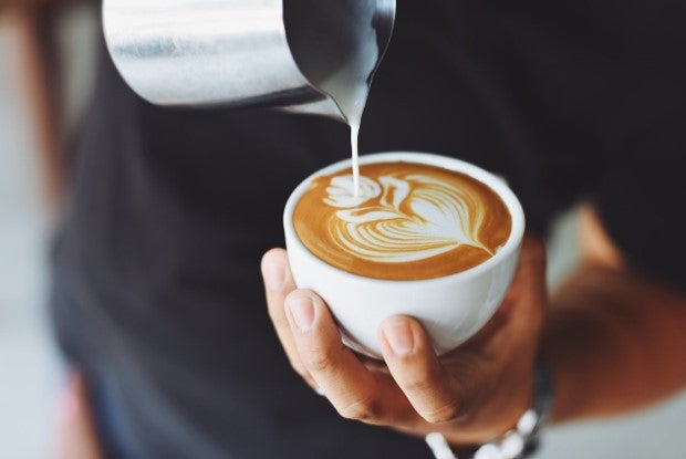 A person pouring milk from a jug into a steaming cup of coffee on a wooden table.