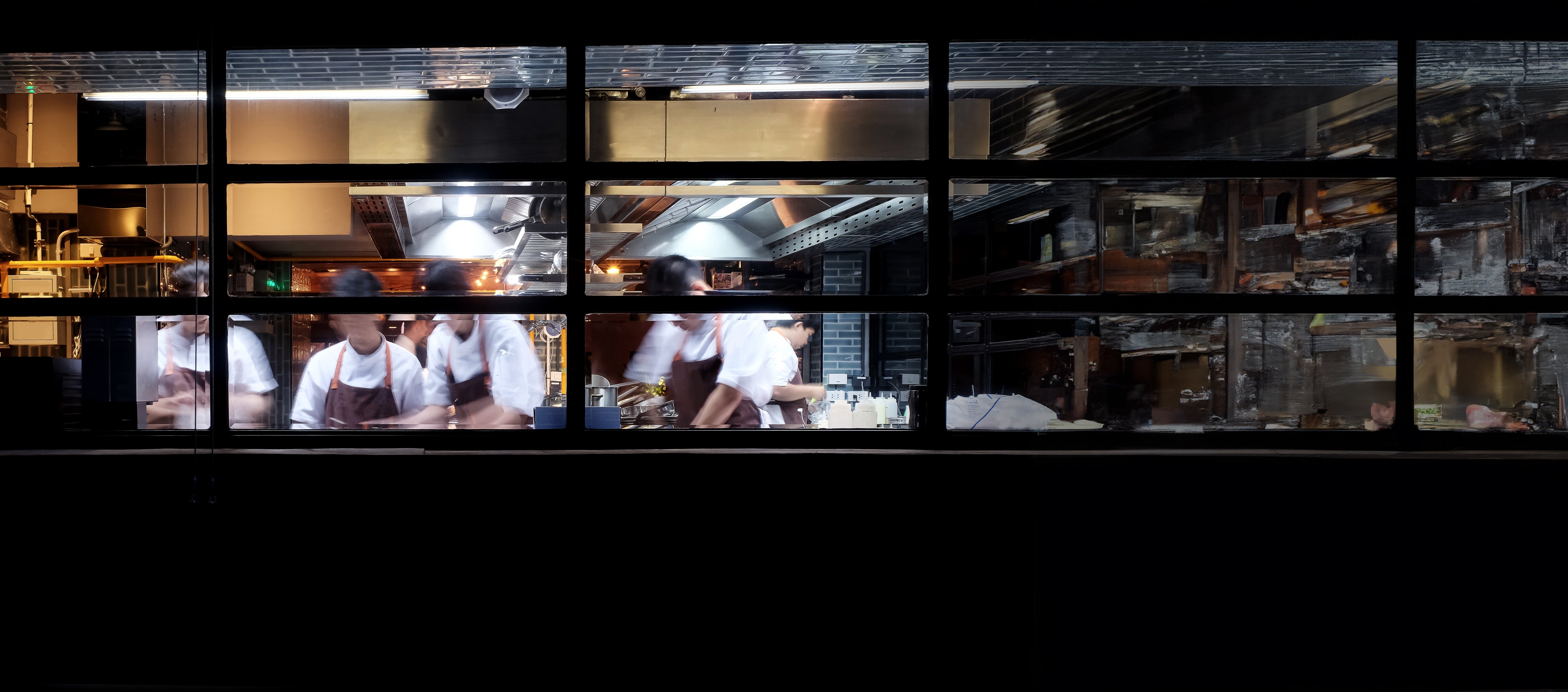 Busy restaurant kitchen seen through window with chefs preparing food in motion blur.