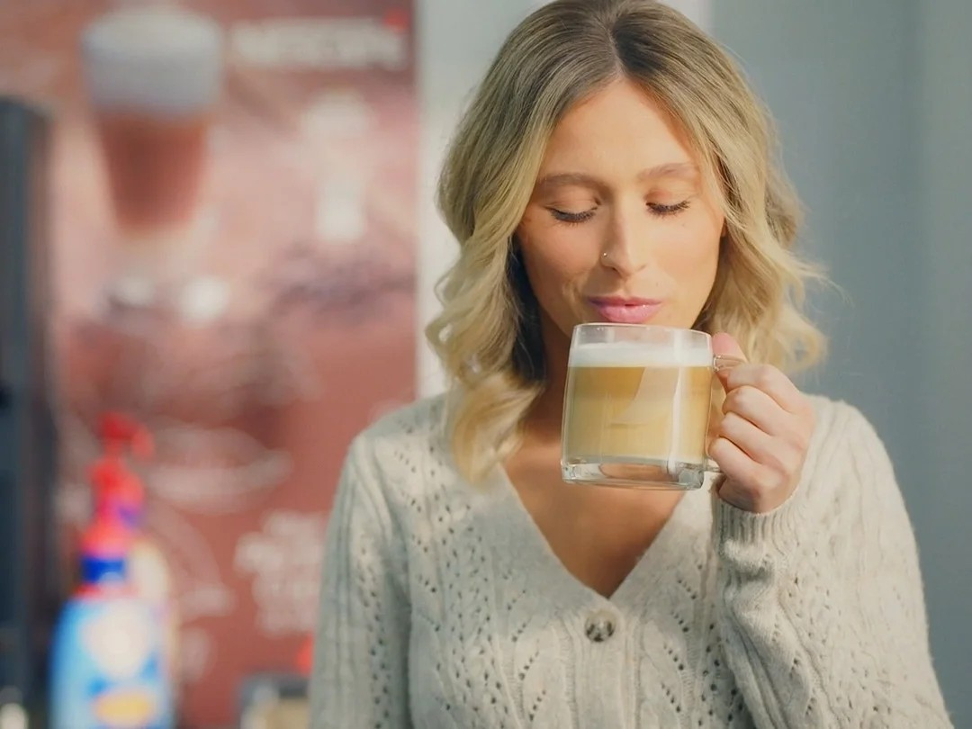A woman holding a cup of coffee stands in front of a coffee machine with various creamers displayed nearby.