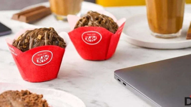 A laptop sits beside a plate of colorful cupcakes and a steaming cup of coffee on a wooden table.