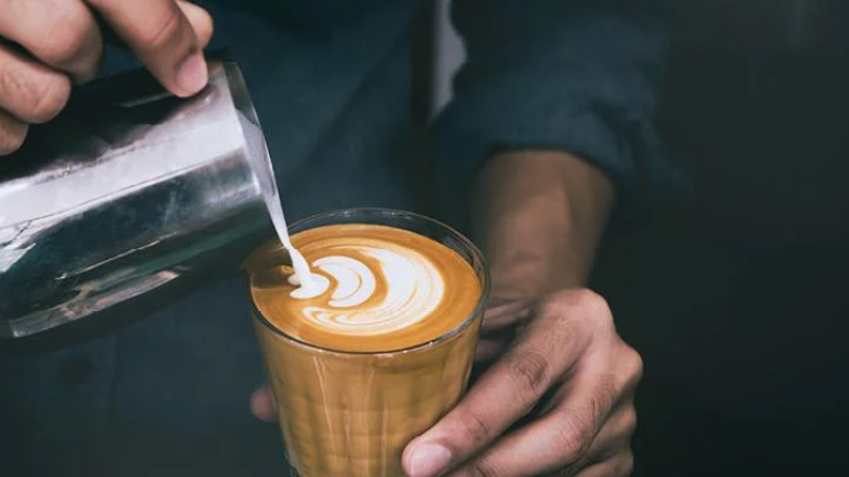 A person pours steaming coffee from a pot into a white cup on a wooden table.