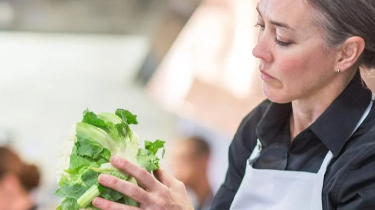 A woman stands in a kitchen, smiling as she holds a fresh salad in her hands.