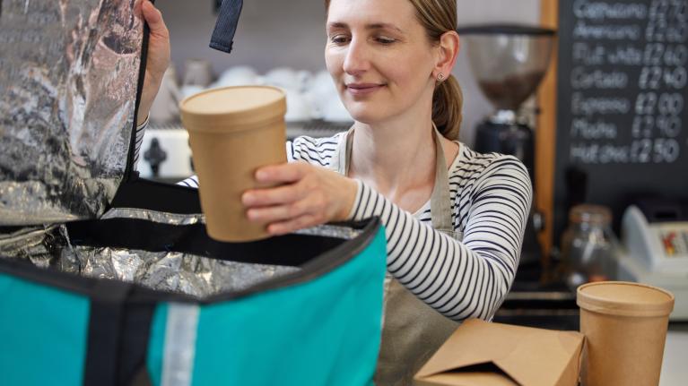 A woman holds a coffee cup while carrying a bag, smiling in a casual outdoor setting.