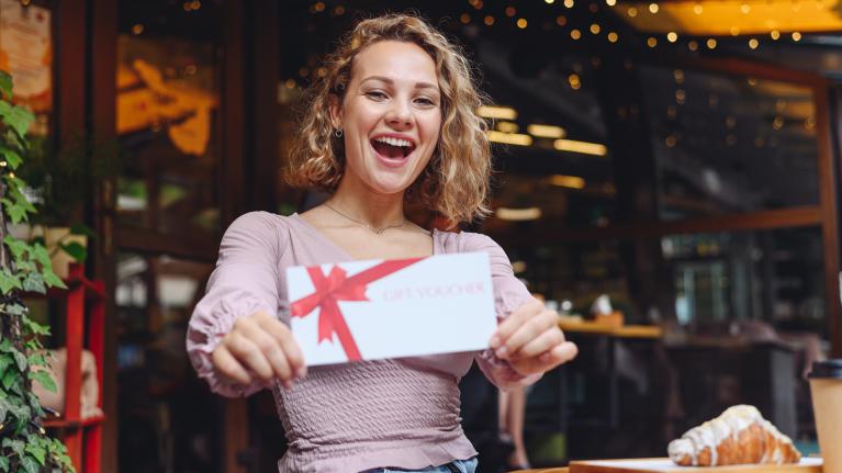 A woman joyfully holds up a gift card, showcasing her excitement and anticipation for the gift inside.