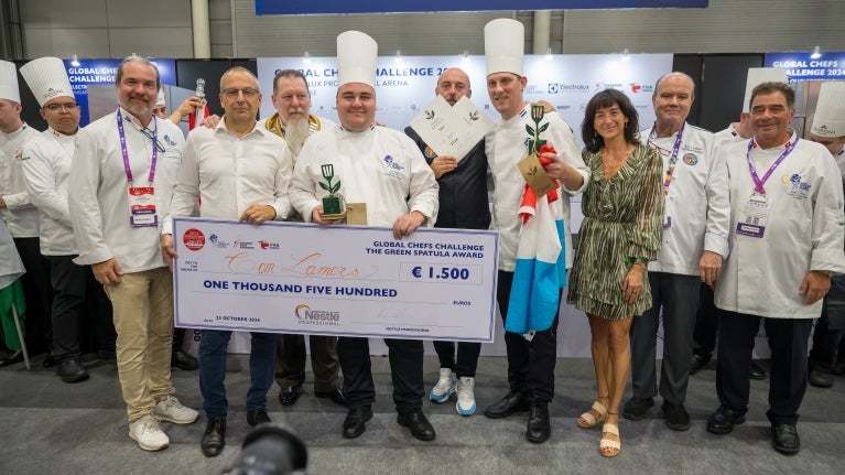 A group of smiling chefs stands together, holding a large check in celebration of their culinary achievement.