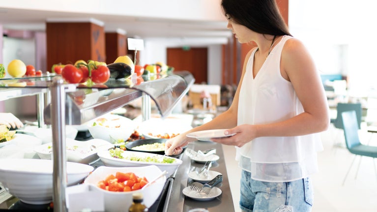 Woman choosing salad at a restaurant buffet counter with bowls of fresh vegetables.