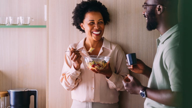 Smiling woman eats salad from a glass bowl while chatting with a man holding a cup in a workplace kitchen.