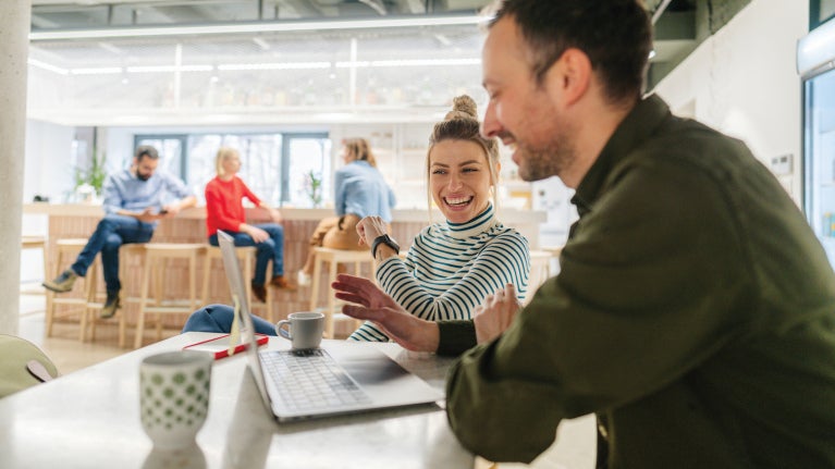 Two coworkers smiling and collaborating over a laptop in a bright, open office.