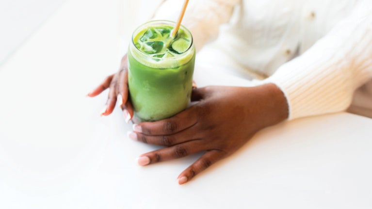 Hands holding a matcha drink with a straw on a white table.