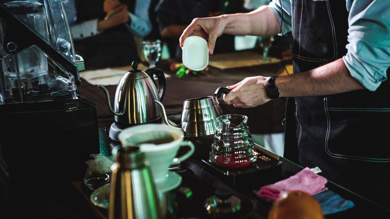 A man in a black apron pours coffee from a pot into a white cup, showcasing a cozy café scene.
