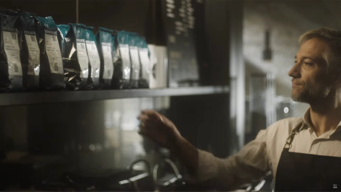 Barista reaching for coffee bags on a shelf in a dimly lit modern café setting.