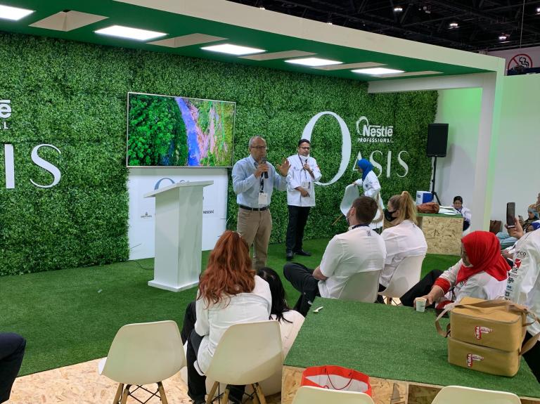 People sitting at tables, chatting and laughing, with a green wall serving as their backdrop.