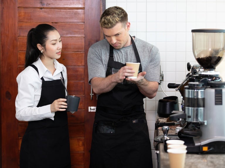 A man and woman stand together, smiling in front of a coffee machine in a cozy kitchen setting.