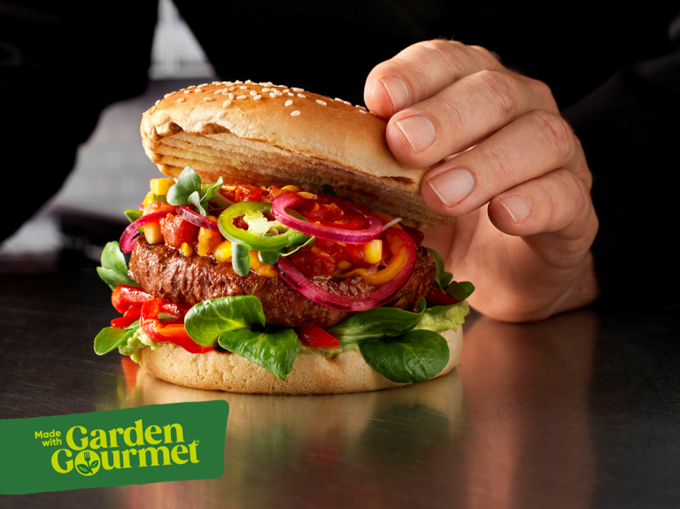 A person holding a burger topped with a fresh green leaf, showcasing a delicious meal option.