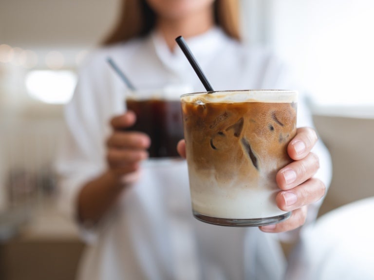 A woman smiling while holding two glasses of iced coffee, showcasing a refreshing beverage choice on a sunny day.
