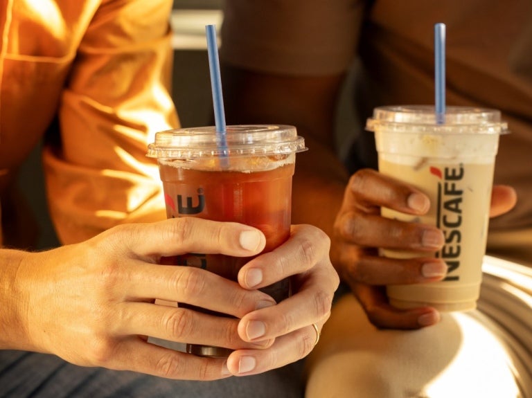 Two people sit on a bench, each holding a Nescafé drink and enjoying a casual conversation.