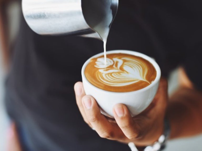 A person pouring milk from a jug into a steaming cup of coffee on a wooden table.