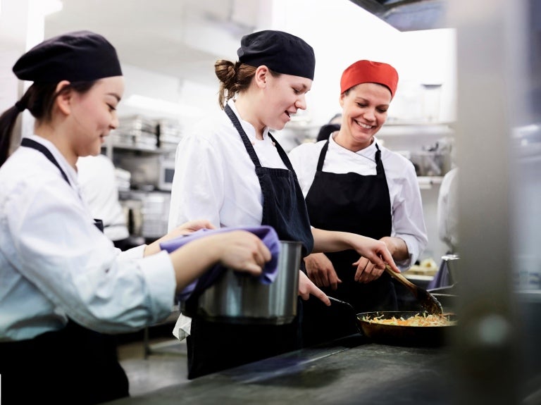 Chefs working together in a busy commercial kitchen, chopping vegetables and cooking on stovetops.