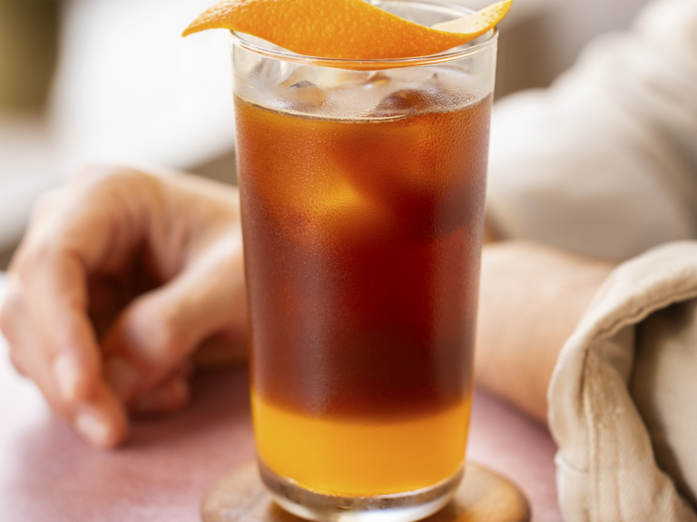 A person holding a tall glass of orange iced tea with ice cubes and a slice of orange on the rim.